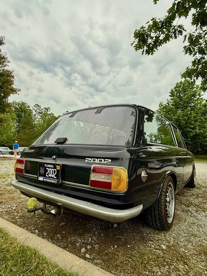 Rear three-quarter view of a black classic BMW parked beside trees under a cloudy sky.