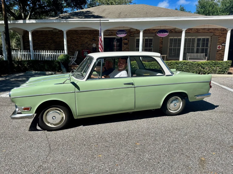 Pale green classic BMW with occupants inside parked in front of a porch-fronted building.