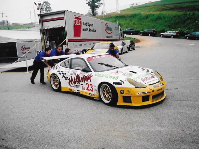Crew members pushing a white and yellow number 23 race car beside a transport truck.