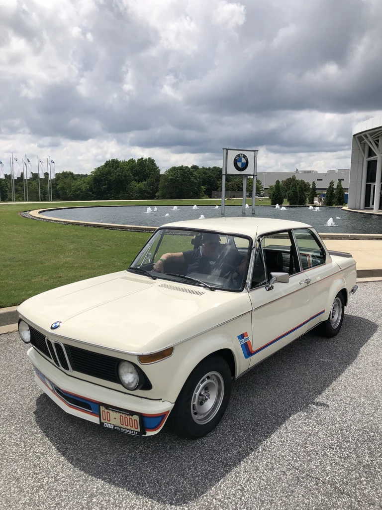 Front three-quarter view of a cream-colored classic BMW beside a fountain and BMW sign.