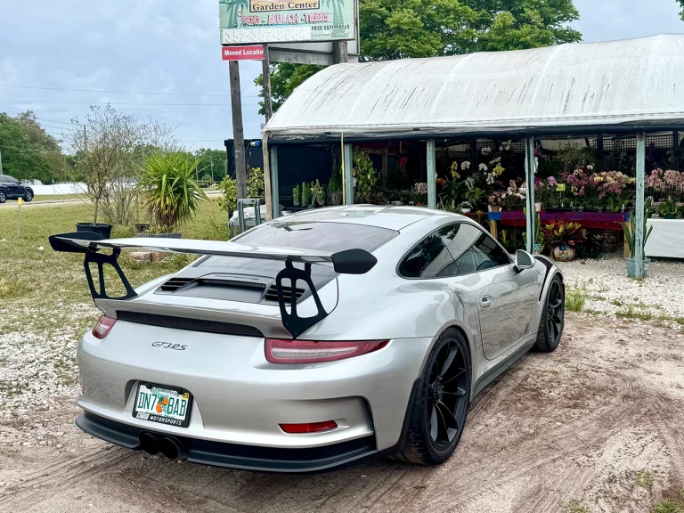 Rear three-quarter view of a silver Porsche with a large rear wing parked outside a garden center.