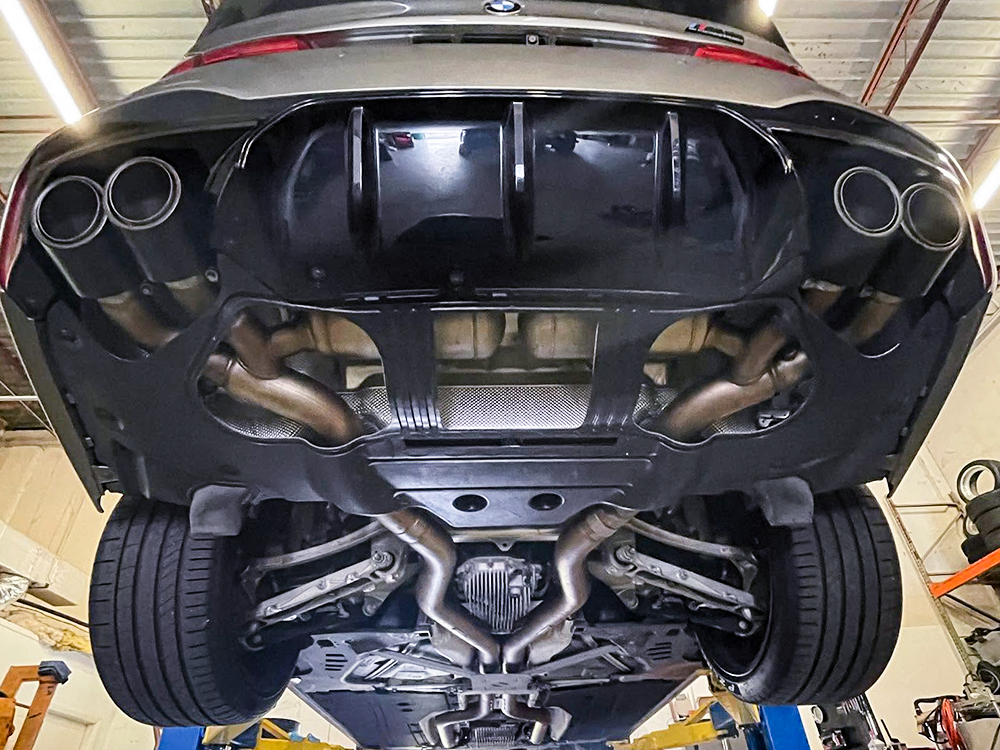 Underside of a BMW on a workshop lift, showing the exhaust system, rear suspension, and quad exhaust tips.